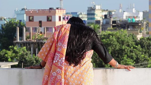 A Bengali woman dressed in a saree stands alone on the rooftop of a house on a bright, sunny day, looking down at the outdoor activity. Her long black hair moves in the gentle breeze. Back view.