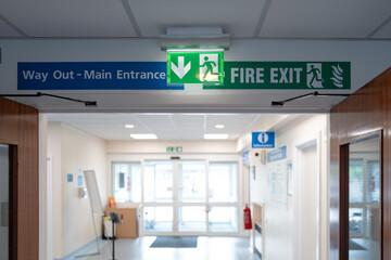 Shallow focus of a typical illuminated Fire Exit sign seen within an NHS hospital outpatients department. The fairy exit leads to the car park and the AE department.