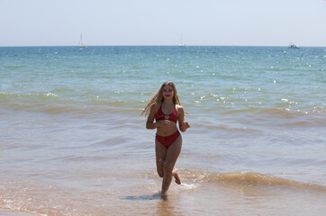 Teenage latin woman, Latin, blonde, young girl runs off the beach between the waves of the sea on the shore. The girl is wearing a metallic red bikini and is on vacation in andalusia, Spain.