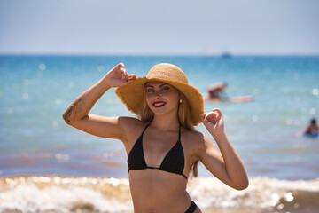 Latin woman, blonde, young and beautiful with blue eyes and black bikini on the shore of the beach with straw hat protecting herself from the sun. In the background the blue sea and a sailboat.