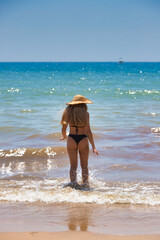 Latin woman, blonde, young and beautiful with blue eyes and black bikini on the shore of the beach with her back turned and wearing a straw hat. In the background the blue sea and a sailboat.