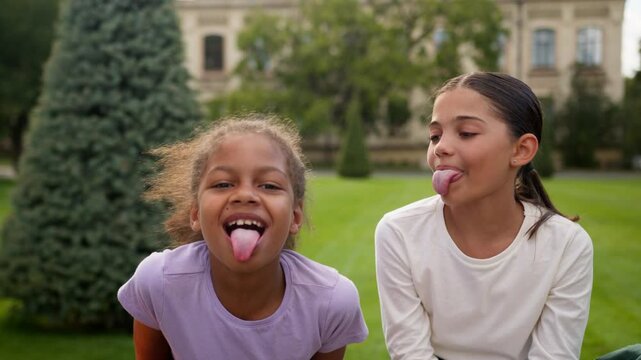 Funny happy girls two little children kids female friends pupils school education multiracial african arabian schoolgirls students sisters daughters stick out tongue grimace play game fun outdoor city