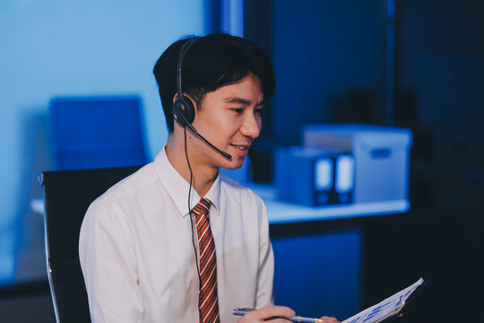 Happy smiling Asian man work at call center service desk consultant with teammates at night, customer service executive with microphone headset use computer for supporting in late night office.