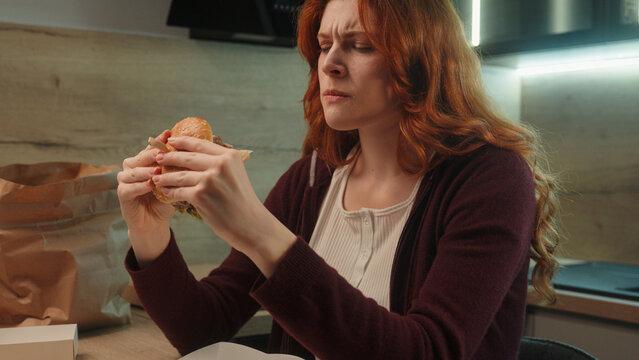 Unhappy displeased Caucasian woman hungry female girl upset lady client customer eating unappetizing cheeseburger chewing disgusting burger taste hamburger junk fast food quality order delivery indoor