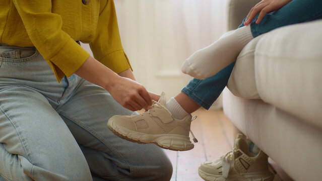 Unrecognizable loving Caucasian woman mother carer female hands lacing ties shoelaces unknown little girl daughter helping tying sneakers child kid in living room at morning. Upbringing family support
