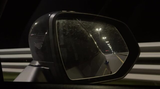 The reflection of night sky and road in rearview mirror of a car driving down a dark highway. Image is captured from right rear view mirror. Modern mirror of black car reflects passing street lights.