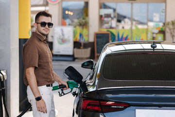 Fototapeta premium Handsome man in sunglasses refuelling his car on self service gas station. Male filling diesel at gasoline fuel and look into camera