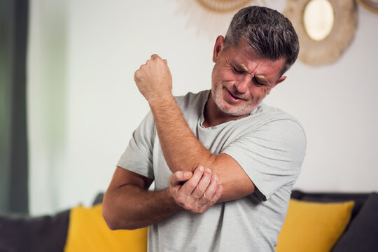 Man With Pain In Elbow. Pain relief concept. Middle age man feeling the pain in hand elbow at home. Male touching his painful elbow