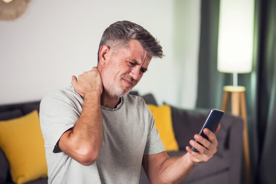 Man with a beard sitting in a living room, touching his neck in discomfort, while holding a smartphone.