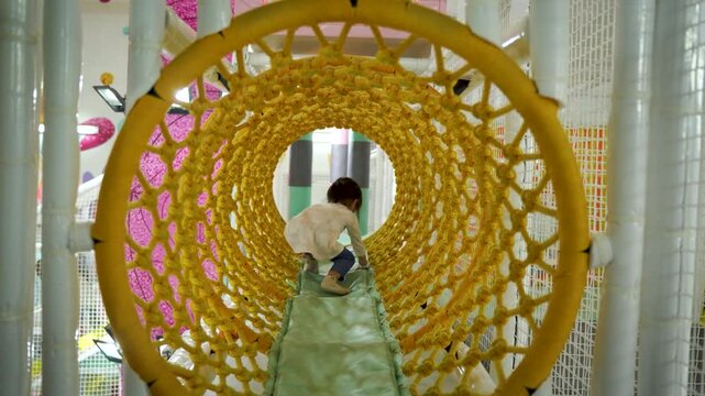 A cute little girl is enjoying her playtime in an indoor playground, entering the net tunnel slowly and carefully.