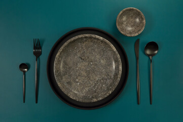 Top view of brown marble plate and bowl, black plate and cutlery on dark green background. Food...