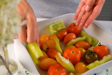 Woman hands adding vegetables pepper tomatoes to baking sheet with oil, garlic for bake vegetables. Female preparing dish on kitchen at home. Ingredient recipe cuisine culinary. Homemade healthy meal.