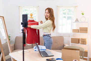 A young woman is showcasing a red sweater on a mannequin while filming content for her online clothing business. She is smiling and organizing her workspace in a bright, well-lit room