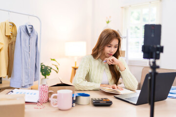 A young woman is sitting at her desk, working on her laptop while filming content for her online clothing business. She is surrounded by clothing, a mannequin, and packaging supplies.