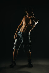 Shirtless athletic man posing in dark lighting wearing ripped denim shorts, showcasing his muscular physique for a fitness and fashion photo shoot.