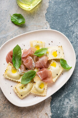 Beige plate with ravioli, prosciutto and fresh basil, vertical shot on a beige and grey granite background, elevated view