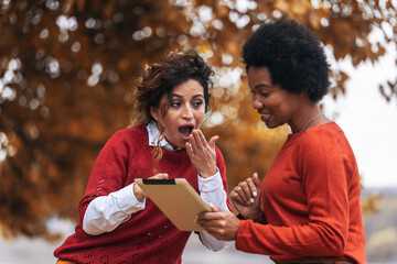 Naklejka premium Two happy female friend talking and sharing life stories, enjoying autumn outdoors, using digital tablet and phone. Women's friendship
