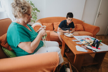 Obraz premium A grandmother sits on a sofa knitting a vest while her young grandson enjoys his meal at the table. The cozy scene captures family bonding moments.