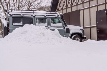 A jeep parked in winter near a country house. The car is parked in a snowdrift next to the house. The snowfall covered the car. It's cold in the village in winter.