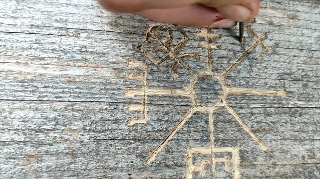 A man's hand with a knife carves the Viking rune of the Vegvisir compass on hard wood, close-up