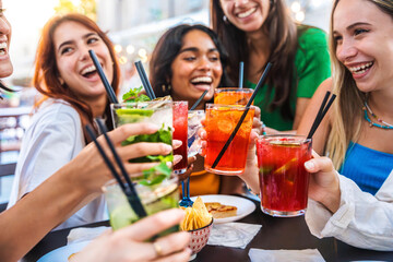 Happy female people toasting cocktail glasses sitting at bar table - Cheerful friends enjoying happy hour at pub restaurant balcony - Food and beverage life style concept