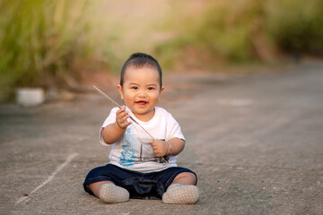 cute asian baby sitting on the road. baby boy holding wild grass while smiling sweetly wearing a white shirt towards the camera. photo atmosphere in the afternoon.