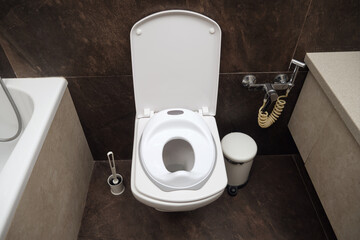 Open white toilet with child potty seat attachment in a bathroom. Bathroom cleaning brush, trash...