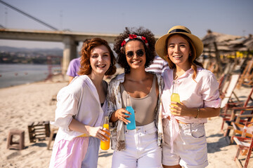 Three girl friends met at the beach to hang out. They are drinking cocktails and walking on the sand.