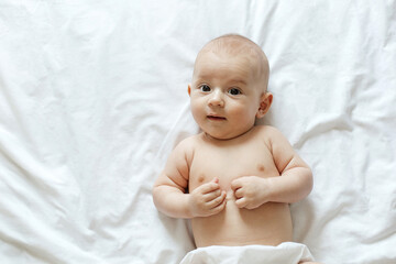 A cheerful baby is lying on a soft white blanket, engaging with the surroundings. The little one displays curiosity, resting comfortably and radiating joy.