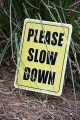 A black and yellow sign that says Please Slow Down, tucked in some green grass.
