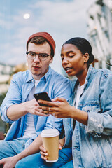 African american young woman showing app on smartphone wondered hipster guy dressed in casual denim wear sitting outdoors with coffee.Multicultural friends watching video on website on cellular