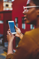 Rear view of african american woman holding smartphone connected to 4G internet standing on street, selective focus on modern mobile phone in hand of dark skinned hipster girl  on urban setting