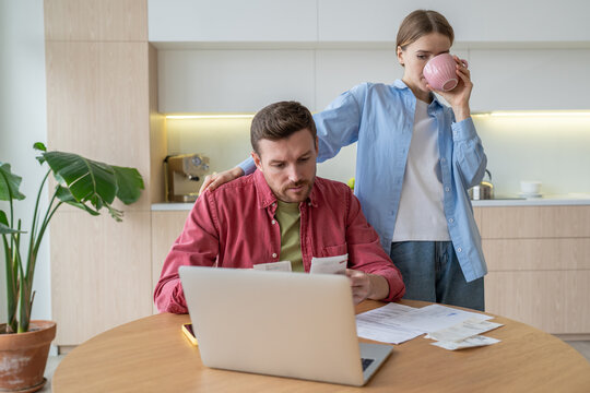 Husband busy with bill calculations, taxes, rent before laptop, wife sips tea beside. Managing family finances, overdue payment, utility expenses, debt, bankruptcy, spenders, inability to manage money