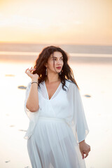  girl with black curly hair in a white dress on the shore of the lake in the sunset light. calm sensual portrait. 