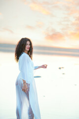  girl with black curly hair in a white dress on the shore of the lake in the sunset light. calm sensual portrait. 