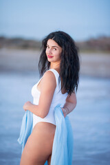 Girl with long curly hair in a bodysuit and shirt walks along the lake shore in the water at sunset.
