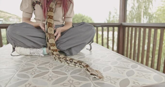 A young woman sits on a patio table gently stroking her pet snake.