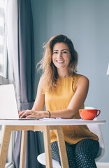 Portrait of happy housewife sitting at table in cozy apartment and spending leisure time for online shopping in web stores, positive female freelancer using home internet connection on laptop