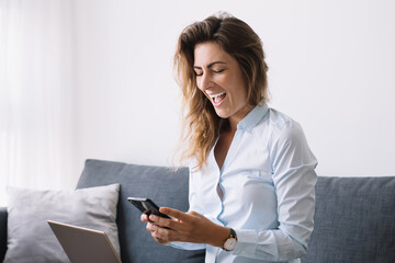 Businesswoman messaging on phone sitting with laptop on knees at home