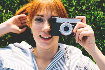 Top view of positive woman photographer in casual shirt lying on summer grass and making pictures...