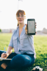 Caucasian woman millennial holding her smartphone with mock up screen showing modern application update for blogging outdoors, selective focus on mobile phone with blank copy space area