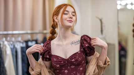 Red-haired woman wearing a floral dress adjusts her jacket in a fitting room. Blurred background with clothes racks. Concept of fashion and self-confidence.