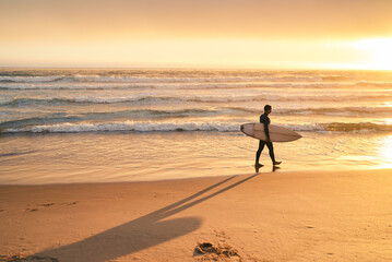 A surfer walking along the beach at sunset, carrying a surfboard. The image captures a serene and peaceful moment.