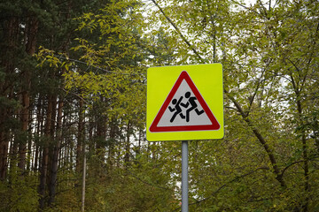Road sign on pole, that is installed near schools. Denoting Be careful, children. Yellow square, red triangle, white, with the image of running people. Safety, Traffic warning, autumn forest.