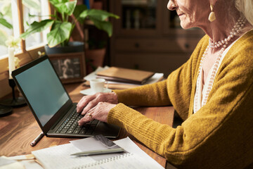 Elderly woman wearing eyeglasses, working on laptop seated at wooden desk, with notebook and coffee...