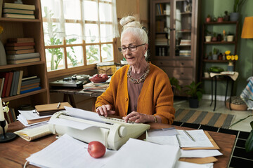 Senior woman sitting at desk filled with papers organizing documents in living room with bookshelves and plants. Natural light coming through window behind providing calm ambience