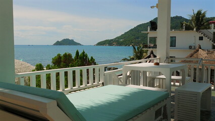Beautiful beachfront terrace with white furniture, overlooking tranquil turquoise sea and lush green hills, ideal for relaxation and vacation, Coastal Living and Serenity.