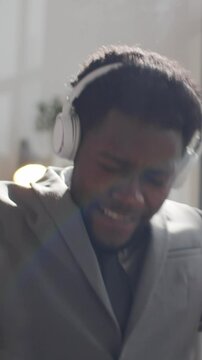 Vertical shot of young Black businessman in formalwear putting on wireless headphones, listening to favourite music, singing and dancing with excitement while sitting at desk in sunlit office