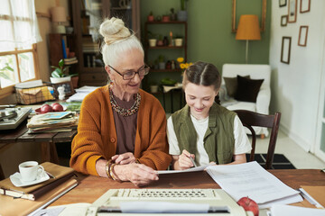 Elderly woman with gray hair and glasses sitting with young child at table, assisting with...