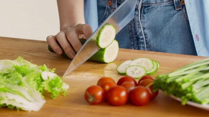 Preparing vegetarian lunch. Woman cutting fresh cucumber for vegetable salad at kitchen table